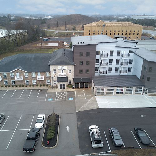 The image shows a drone-view of a contemporary apartment or hotel building with a parking lot in the foreground and a few cars parked.