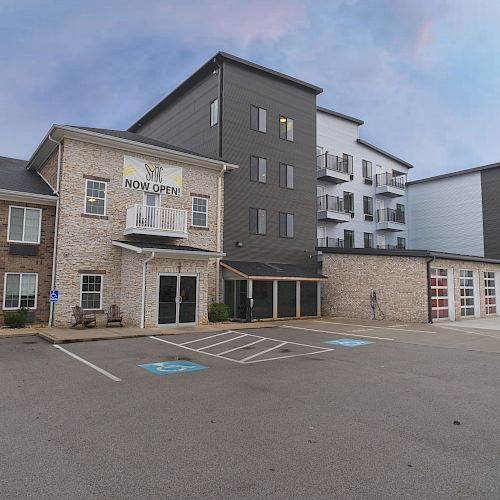 The image shows a modern, multi-story apartment building with a sign indicating it's now open, and a mostly empty parking lot with a few accessible spots.