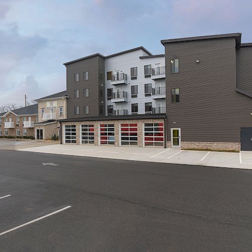 The image shows a multi-story building with garage doors on the lower level, residential balconies above, and an adjacent parking lot in view.