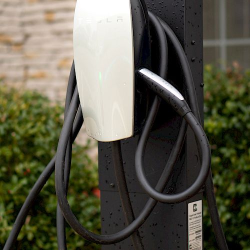 An electric vehicle charging station is pictured with a white charging unit and coiled cables, set against a backdrop of greenery and a stone wall.