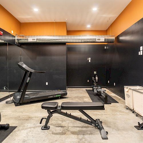 The image shows a modern gym with a treadmill, bench, dumbbells, and exercise machine, black and orange walls and mirrors on one side.