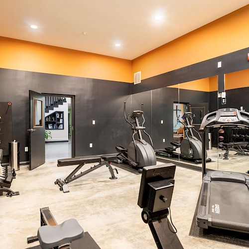 The image shows a gym with exercise machines, including a treadmill, elliptical, and rowing machine. There's a wall mirror and an open door.