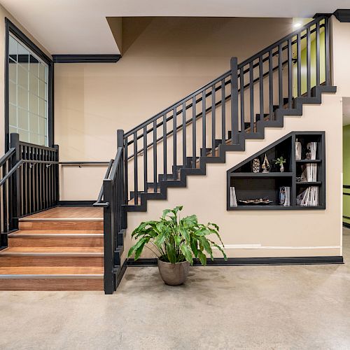 A modern indoor staircase with wooden steps, a plant at the bottom, and a shelf on the adjacent wall, located in a clean, open space.