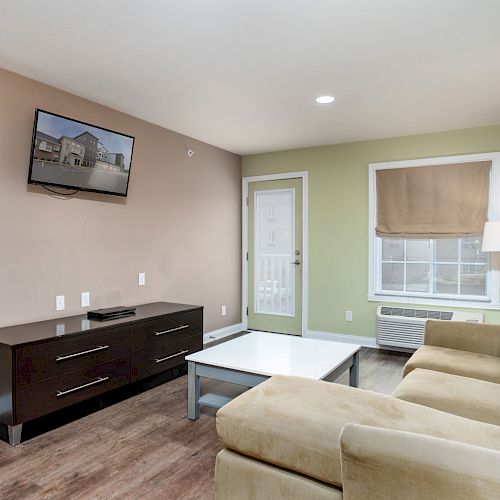 A modern living room with a beige couch, coffee table, TV mounted on the wall, console, lamp, window with blind, wooden floor, and door to the outside.