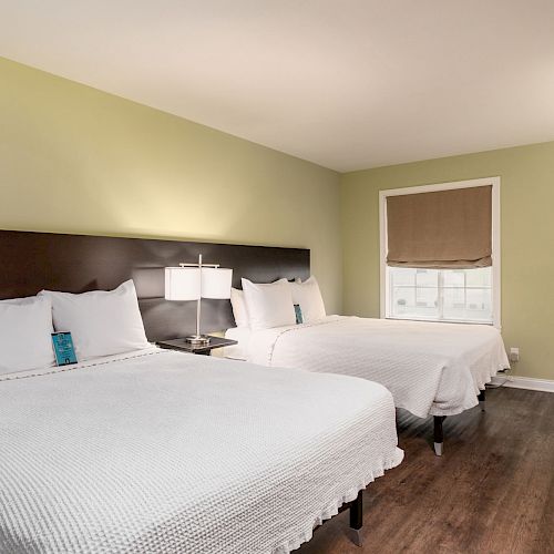 The image shows a hotel room with two double beds, a nightstand with a lamp, a window with a shade, and a desk with a chair.