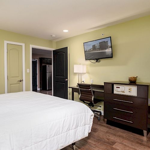 A bedroom with a bed, a desk, a chair, a dresser, and a wall-mounted TV. Walls are light green and the floor is wood.