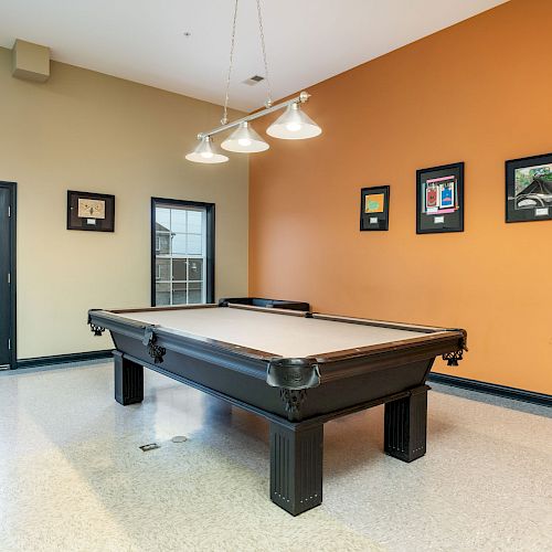 A room with a pool table, orange and beige walls, framed pictures, and a door with a window. The floor is shiny, and there are hanging lights above the table.