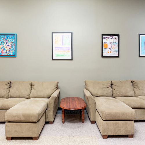 The image shows two beige sofas with chaise lounges, a round wooden table between them, and four colorful framed artworks on the wall above.