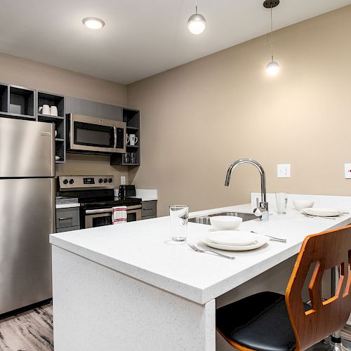 A modern kitchen with a stainless steel fridge, stove, microwave, island with sink, two chairs, plates, and glasses under pendant lights.