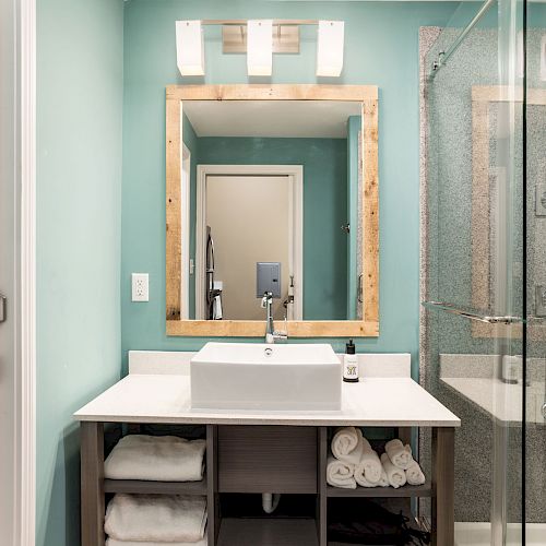 A modern bathroom with a wood-framed mirror, sink, open shelving with towels, a toilet, and a glass-enclosed shower against teal walls.