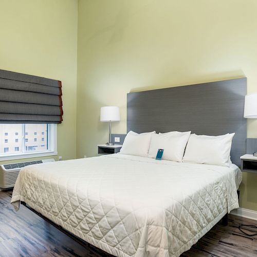 A modern, minimalist bedroom with a large bed, two nightstands with lamps, a window with a Roman shade, and wooden flooring.