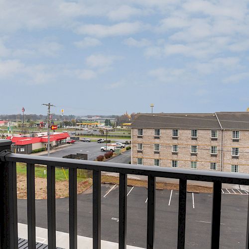 The image shows a black railing balcony overlooking a parking lot, a hotel building, and some businesses including a red-roofed restaurant.