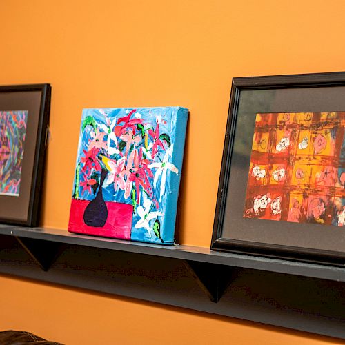 Three colorful paintings on a black shelf against an orange wall. The artworks vary in style and are framed or canvas-mounted.