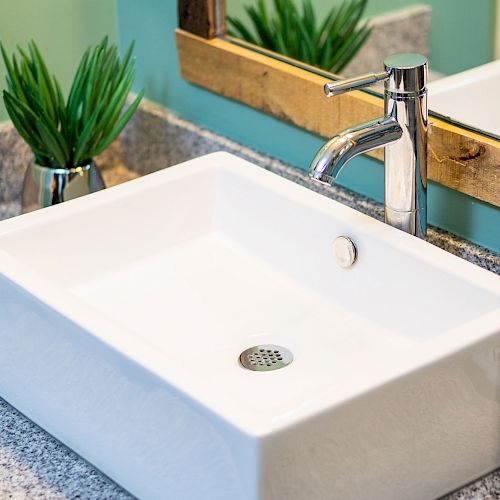 A modern bathroom sink with a rectangular basin, chrome faucet, granite countertop, a mirror, and two small green plants in pots.