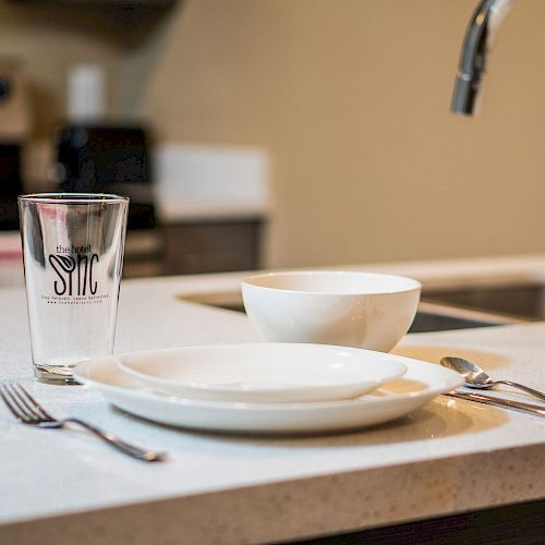 A kitchen counter with a plate, bowl, glass, knife, fork, and spoon, with a sink and a stove in the background.