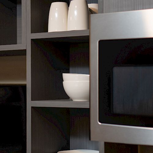 A kitchen shelf holds white plates, mugs, and a bowl next to a microwave, all neatly arranged.