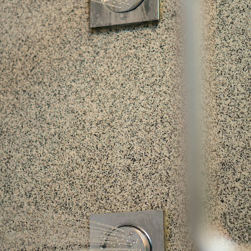 This image shows two square shower jets mounted on a textured wall, with water spraying out, and a blurred object in the foreground.