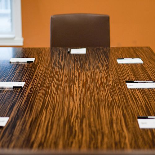 A conference table with six name cards placed on it, surrounded by chairs, in a room with an orange wall and a window in the background.