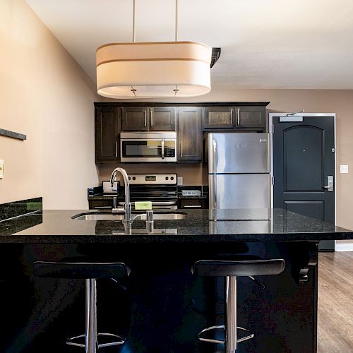 A modern kitchen with a black island, two bar stools, stainless steel appliances, and dark cabinetry. The area has a light fixture above it.