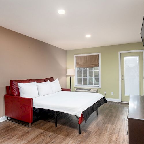 A modern bedroom with a double bed, wall-mounted TV, dresser, lamp, and window. There is a small red bench near the bed and a door leading outside.