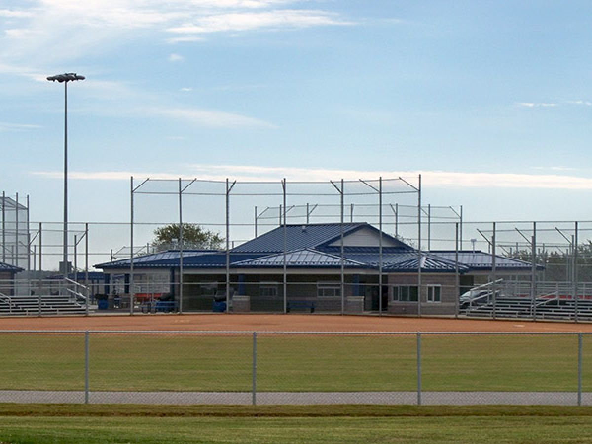 A baseball or softball field with empty bleachers, surrounded by fencing and light towers, under a partly cloudy sky.