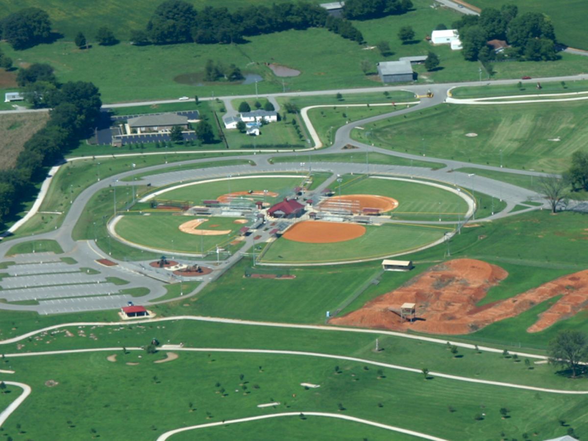 An aerial view of a sports complex with multiple baseball fields, surrounding greenery, and parking areas.