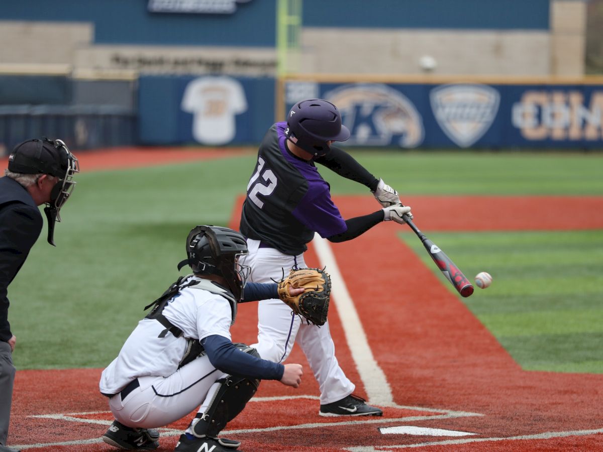 A baseball player in a purple jersey swings at a pitch as the catcher and umpire observe, with a brick building visible in the background.