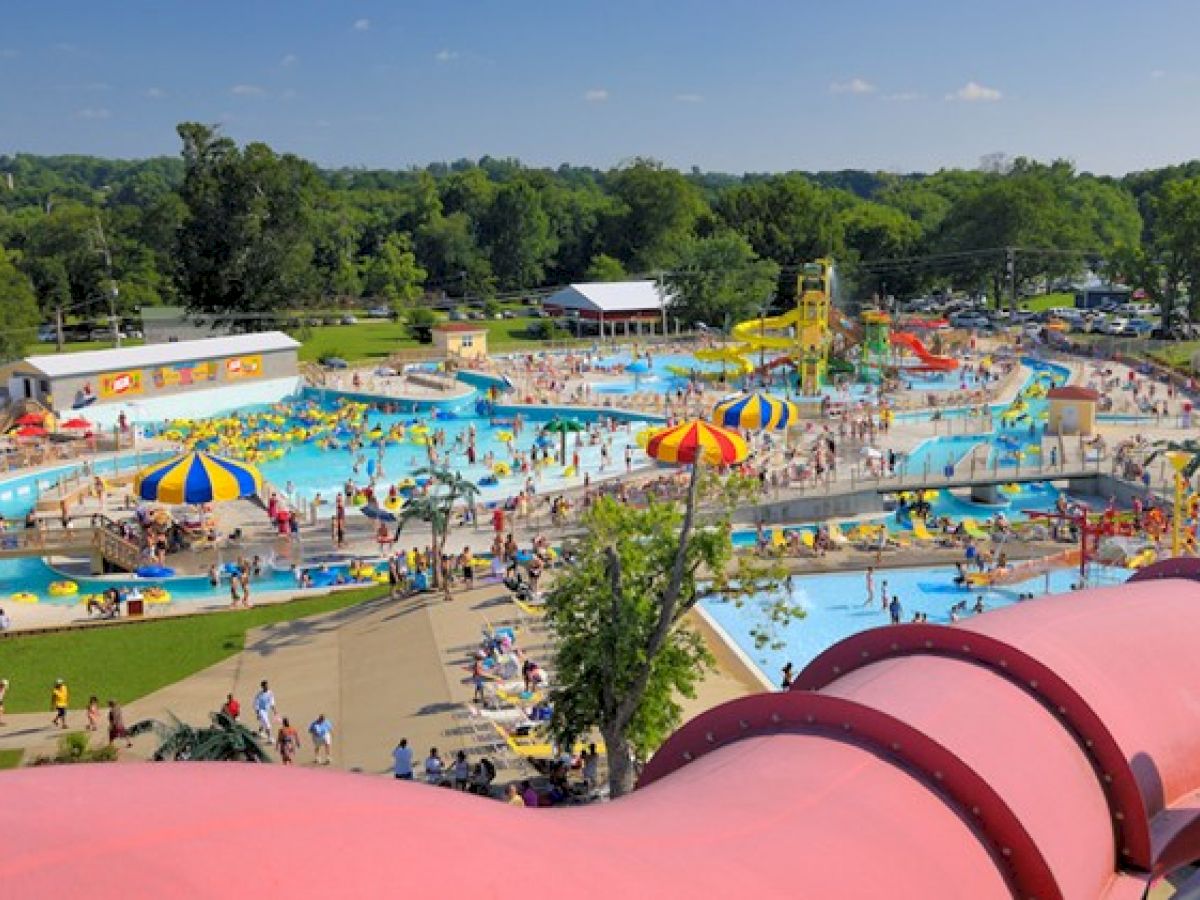 The image shows a bustling water park with various pools, slides, and colorful umbrellas. People are swimming and relaxing, enjoying their day.