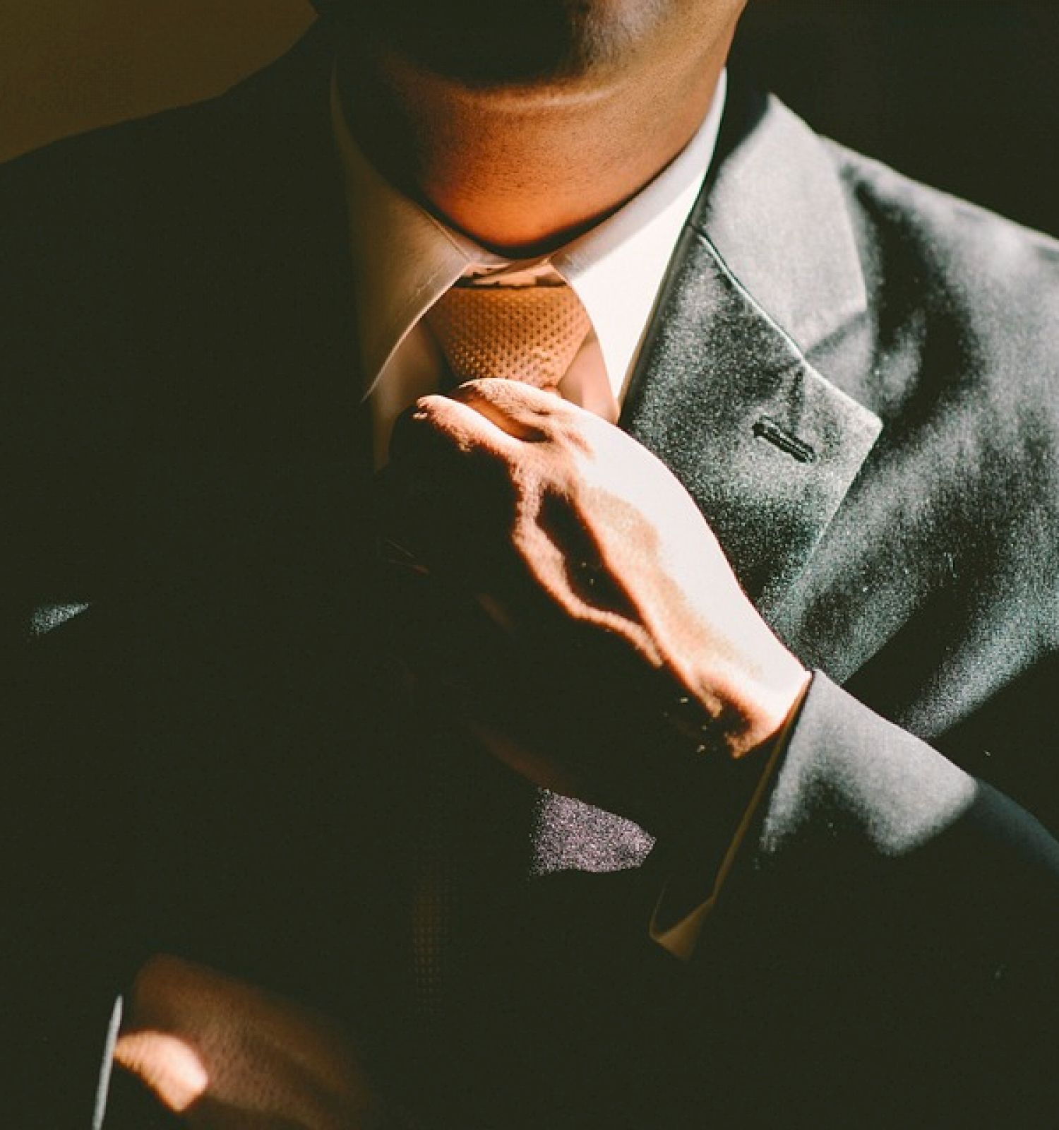 A person in a suit adjusts their tie, with a focus on their hand and upper torso, partially lit by a light source.