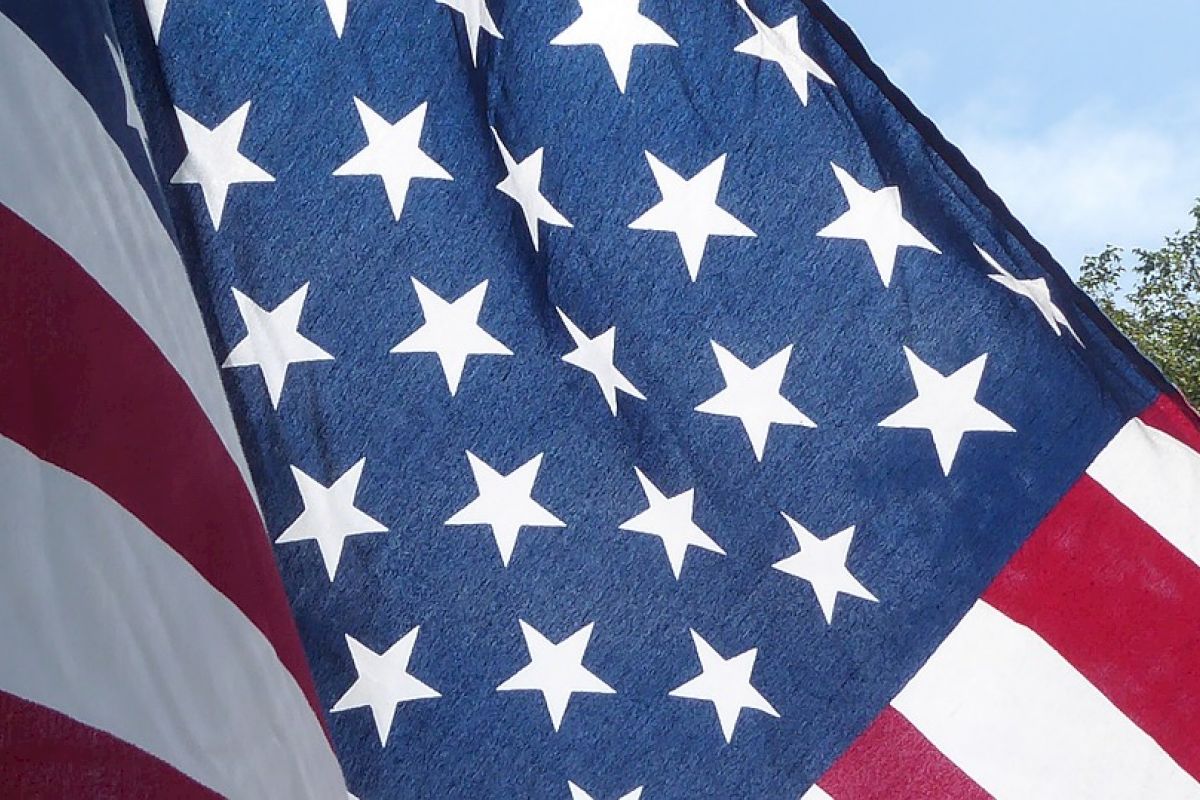 The image shows the American flag, displaying its characteristic stars and stripes, with part of the sky and some greenery visible in the background.