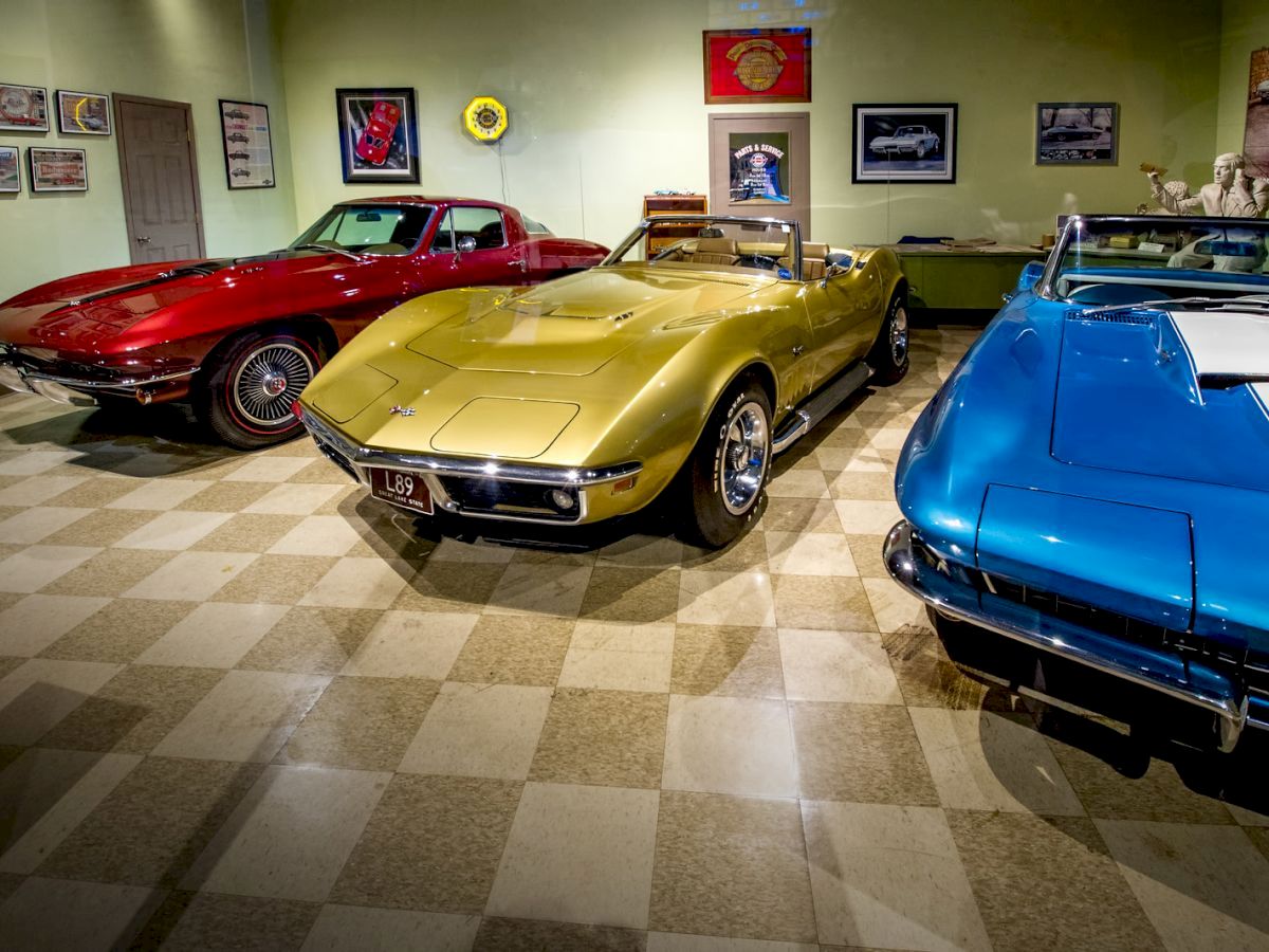 The image shows a collection of classic cars parked indoors; a red, yellow, and blue car are prominently displayed side by side in a well-lit room.