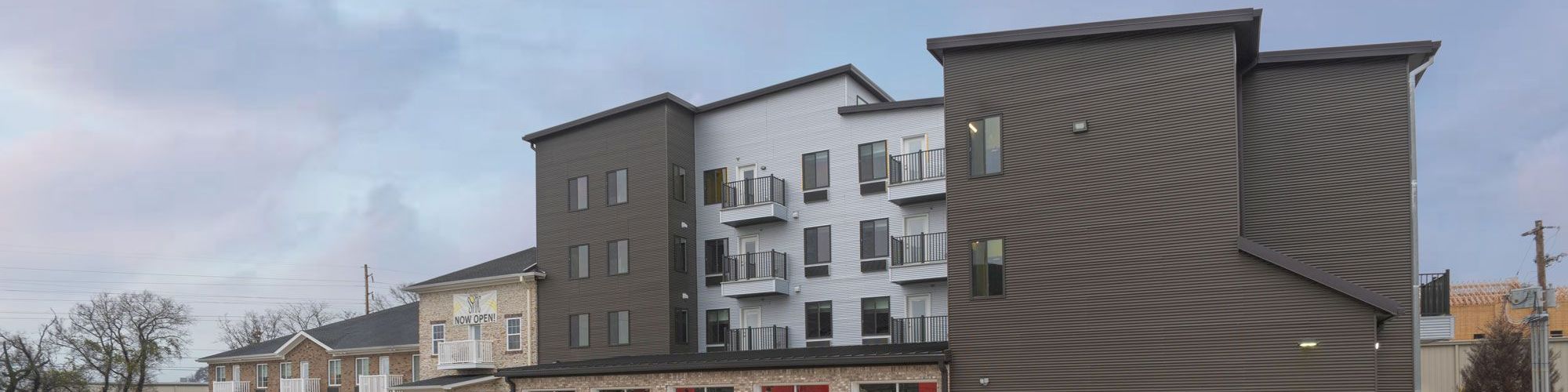 The image shows a modern building with multiple garage doors and residential balconies above, situated next to a large parking lot.