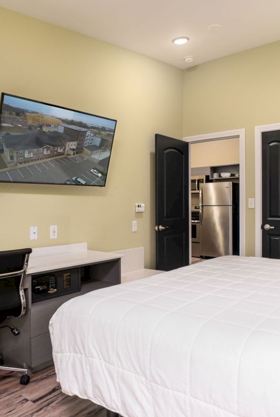 The image shows a modern bedroom with a wall-mounted TV, a desk and chair, and a bed with white linens. There is an open door to a kitchen.