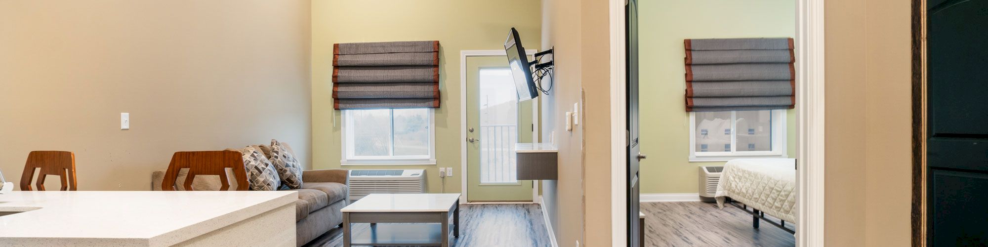 The image shows a modern interior with an open-plan living area, a kitchen island, a wall-mounted TV, and an adjacent bedroom with a neatly made bed.