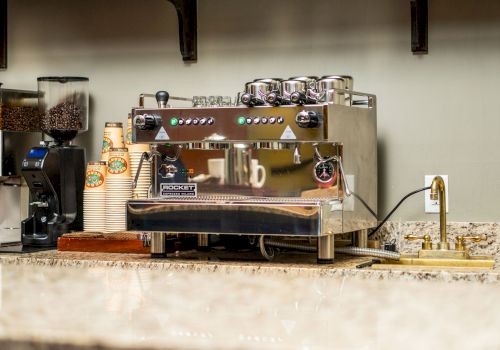 The image shows a coffee shop counter with a large espresso machine, grinders, and a stack of paper cups.