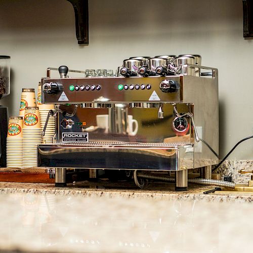 The image shows a coffee shop counter with a large espresso machine, grinders, and a stack of paper cups.