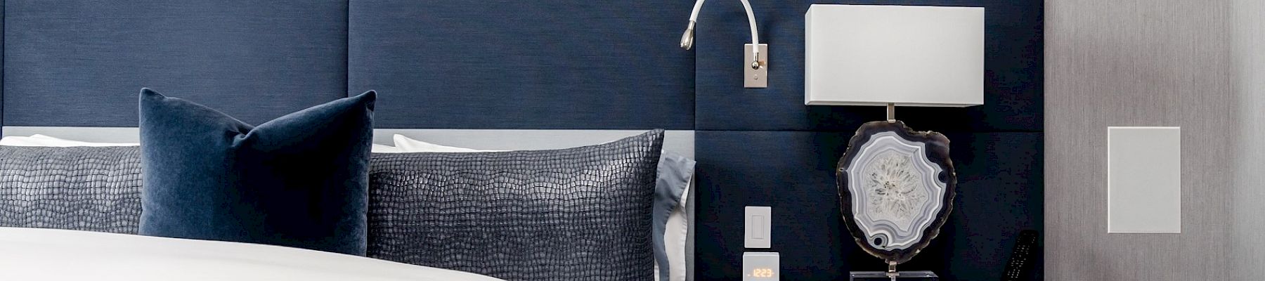 A bedroom with a blue and white color scheme, featuring a bed, nightstand, lamp, and wall-mounted reading light.
