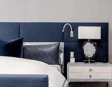 A bedroom with a blue and white color scheme, featuring a bed, nightstand, lamp, and wall-mounted reading light.