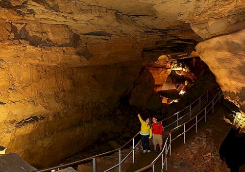 Two people are standing on a pathway inside a large underground cave, with rocky walls and ceiling illuminated by artificial lights.