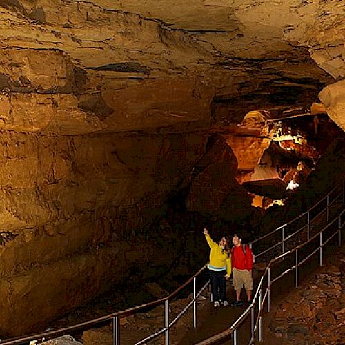 Two people are standing on a pathway inside a large underground cave, with rocky walls and ceiling illuminated by artificial lights.