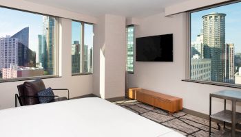 A modern hotel room featuring a bed, a chair, a wall-mounted TV, and a view of a cityscape through multiple large windows.