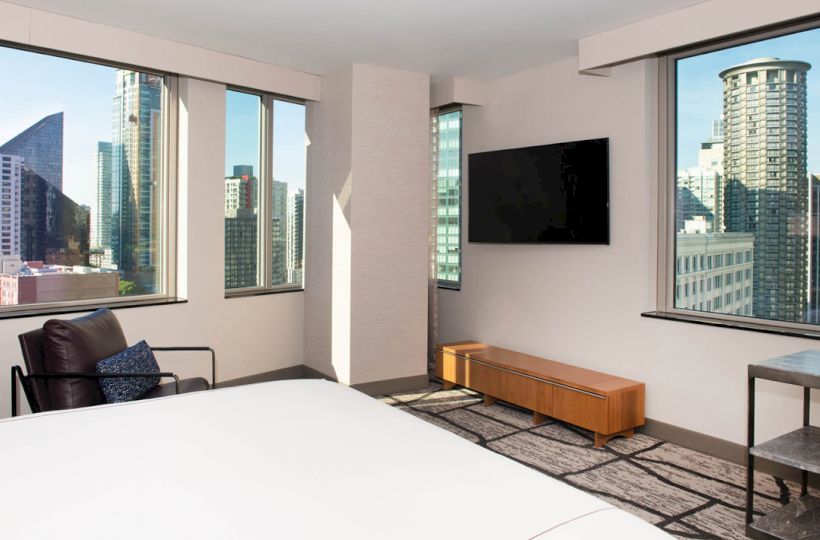 A modern hotel room featuring a bed, a chair, a wall-mounted TV, and a view of a cityscape through multiple large windows.