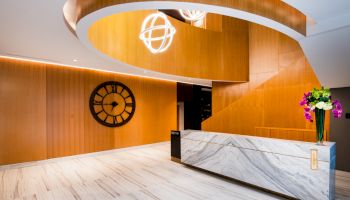 A modern lobby with wooden accents, a large wall clock, a marble reception desk, and decorative lights and flowers.