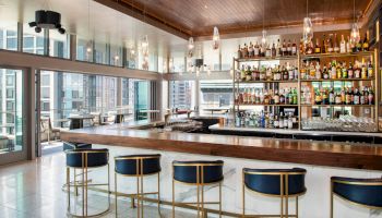 A modern bar with a wooden counter, gold barstools, and a fully stocked liquor shelf in a bright, spacious room with large windows and city views.