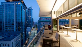 The image shows a modern balcony at dusk overlooking a cityscape with tall buildings. The balcony has seating and tables.