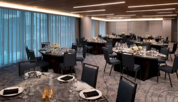 A banquet hall with round tables and blue chairs, set with napkins, plates, and glassware, is prepared for an event.
