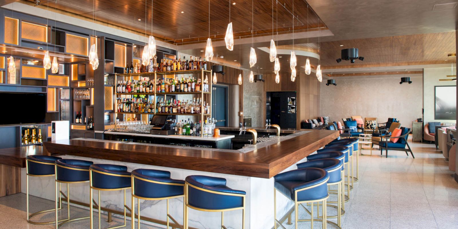 The image shows a modern bar with a long counter, stylish bar stools, hanging lights, and a fully stocked liquor display in a spacious, well-lit room.