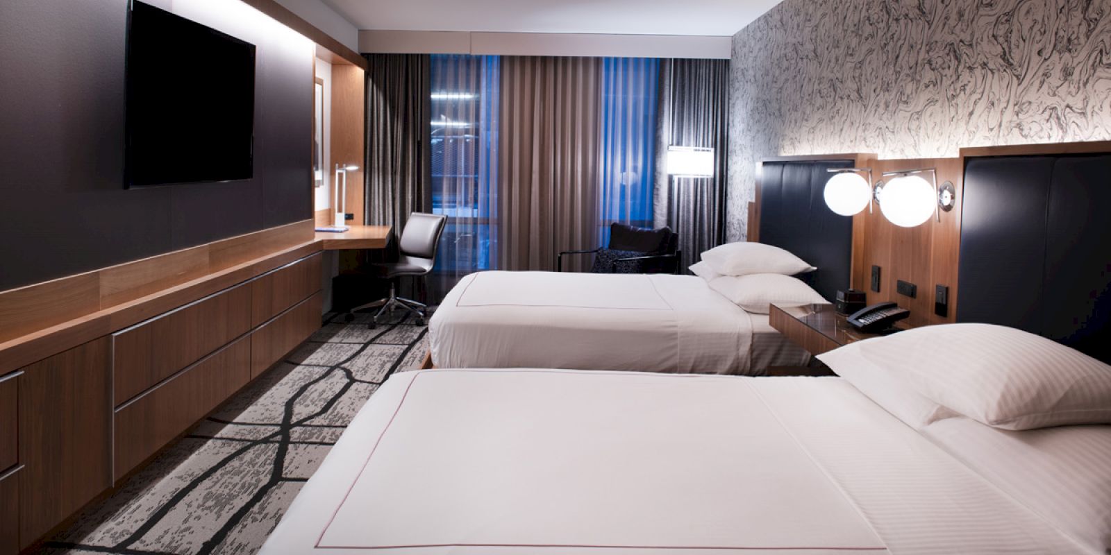 The image shows a modern hotel room with two neatly made beds, a wall-mounted TV, a desk with a chair, and contemporary decor and lighting.