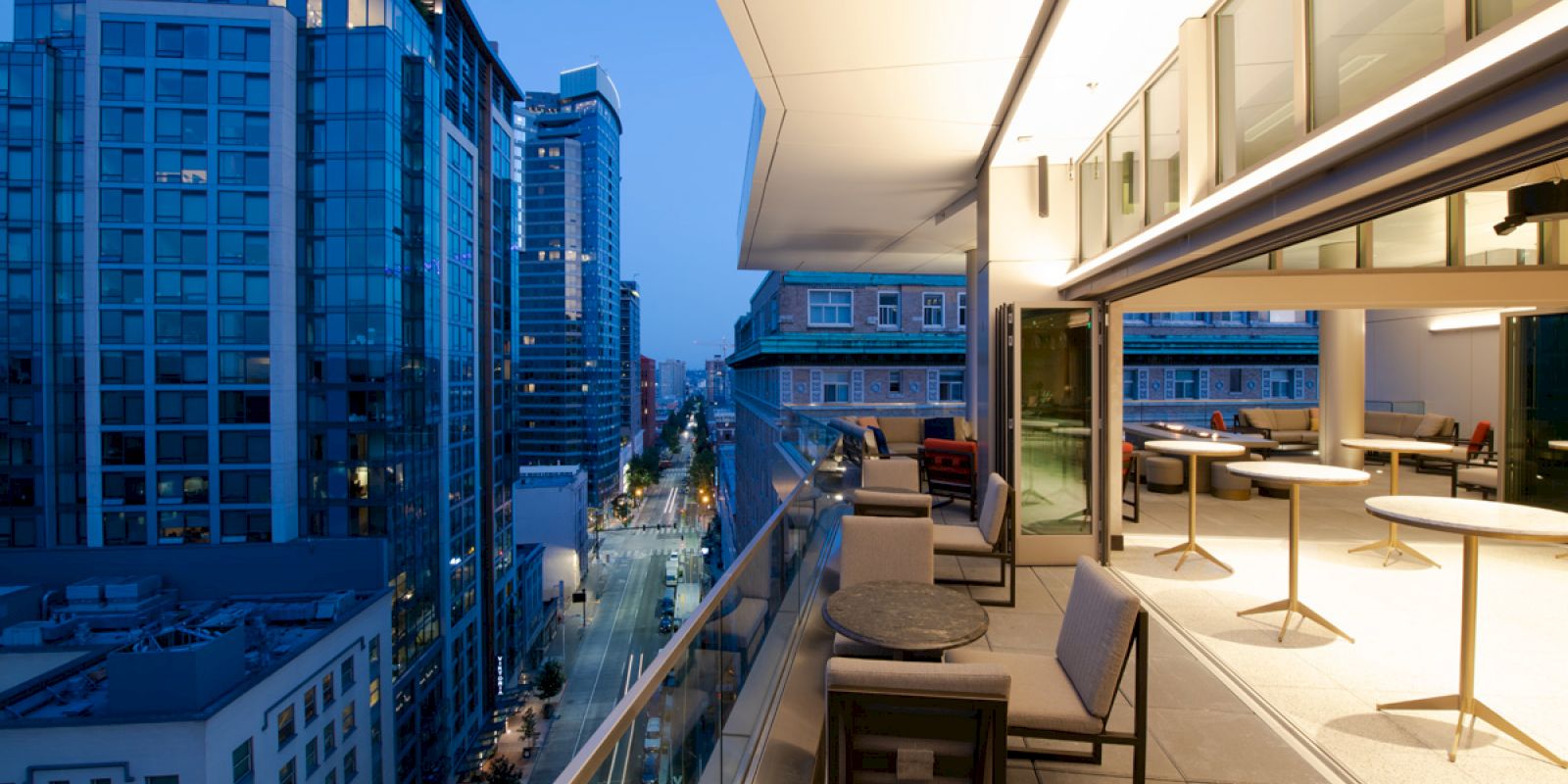 A modern urban balcony with seating areas overlooks a cityscape at dusk, featuring high-rise buildings and a view down a street.