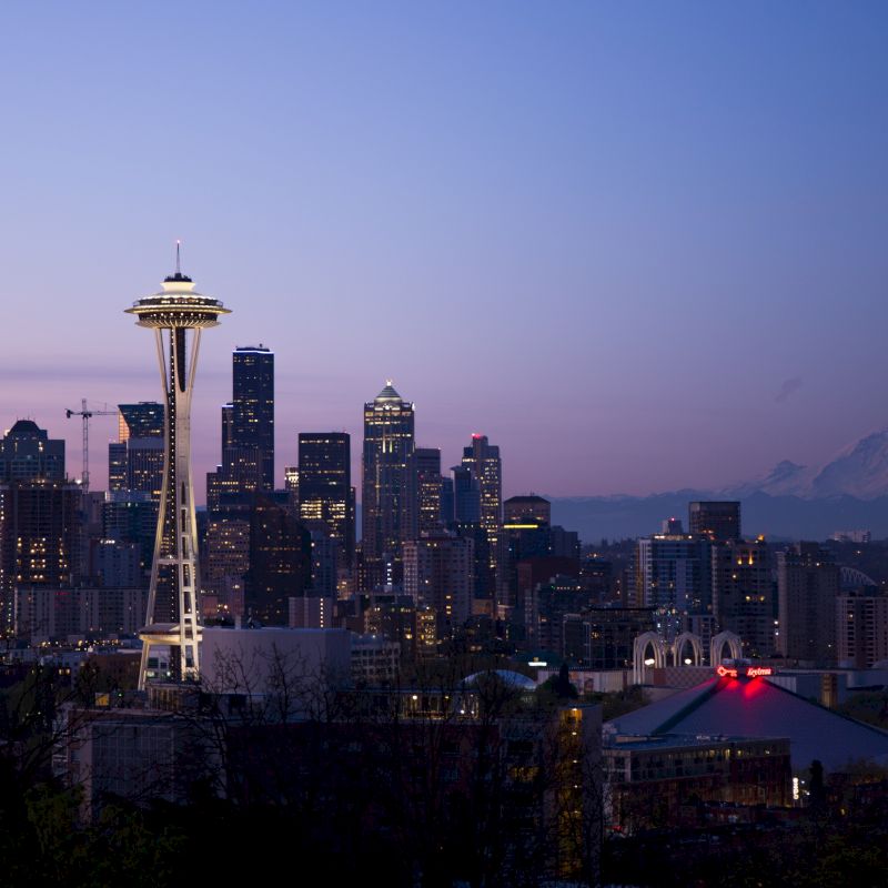 This image shows the Seattle skyline at dusk with the Space Needle prominently featured, Mount Rainier in the background, and city lights beginning to illuminate.